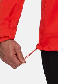 Lys rød jakke med elastiske manchetter og justerbar snor i kanten, med en glat tekstur og en sidelukket lomme.