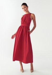 Sieviete sarkān bezpiedurkņu midi kleitā ar savilktu jostasvietu un sānu kabatām, uzvelkusi bēšas, dekorētas, smailas kurpes, stāvot pret baltu fona.