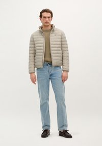 Light grey quilted jacket with a high collar, worn over a green sweater, paired with light blue jeans and dark shoes. Simple design, no patterns.