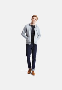 Felpa grigia chiara con zip e tasca frontale, indossata sopra una maglietta nera. Abbinate a jeans blu scuro e stivali marroni con lacci.
