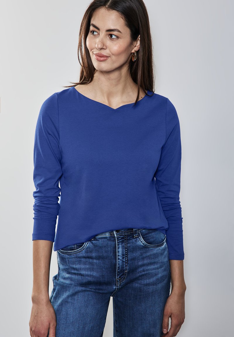 Haut bleu à manches longues avec un col dégagé, fabriqué en tissu lisse. Associé à un jean en denim ajusté, mettant en avant un style décontracté.
