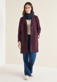 Manteau bordeaux au design ajusté, doté de deux boutons et de poches latérales, associé à une écharpe bleu marine, un haut gris et un jean à jambes larges. Baskets blanches.