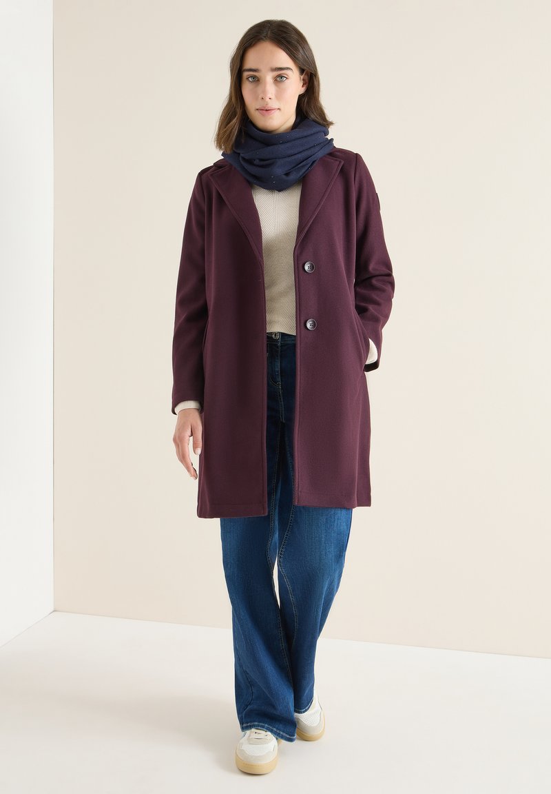 Manteau bordeaux au design ajusté, doté de deux boutons et de poches latérales, associé à une écharpe bleu marine, un haut gris et un jean à jambes larges. Baskets blanches.