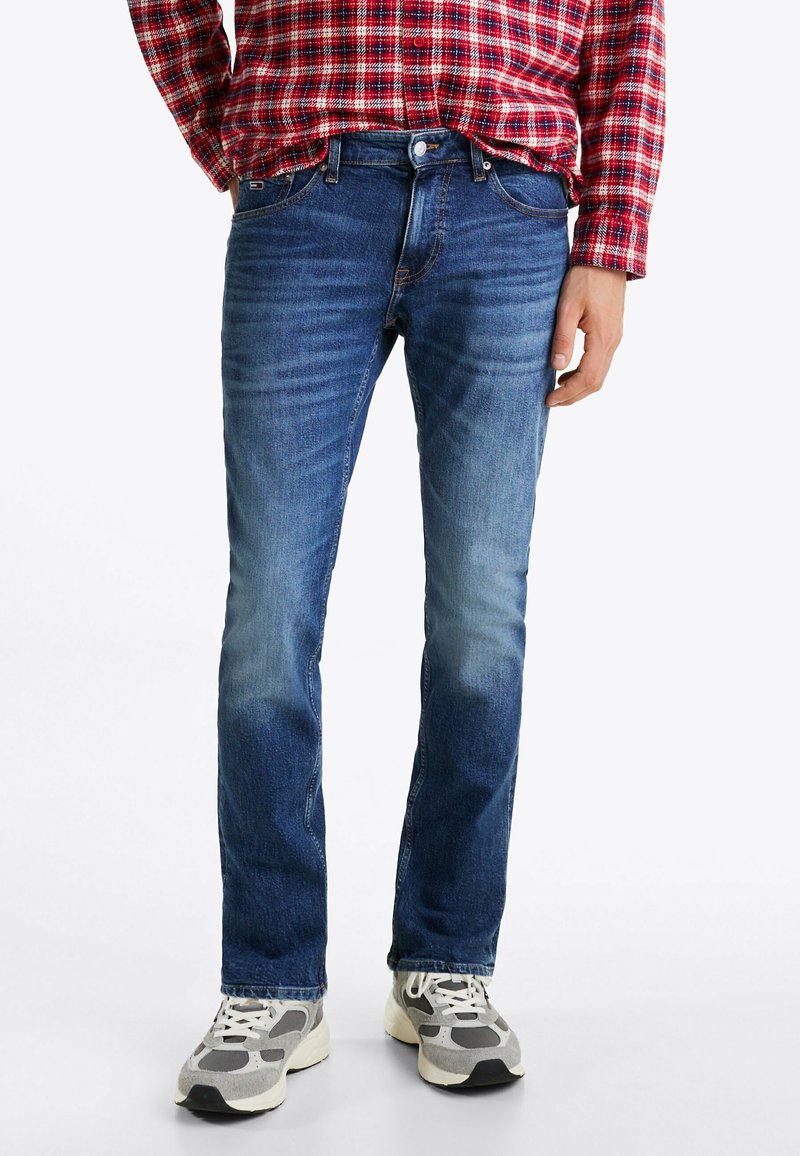 Partie inférieure du corps d'une personne portant un jean bleu délavé, des baskets grises et blanches, et une chemise rouge à carreaux à manches longues.