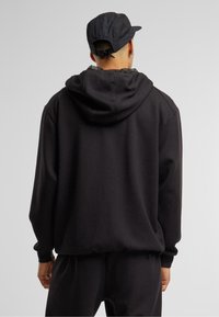 Sudadera negra con un corte relajado, que presenta una gran capucha, puños y cintura acanalados. Textura suave. Vista desde la espalda.