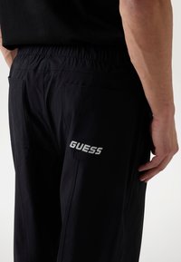 Black cargo pants made from textured fabric. Features include an elastic waistband, side pockets, and embroidered 'GUESS' logo in white.