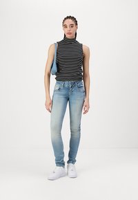 Top sin mangas a rayas blanco y negro, jeans ajustados azul claro y zapatillas blancas. La modelo sostiene un pequeño bolso azul.