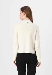 Pull blanc en tricot avec un col haut. Présente une bordure en dentelle florale sur les manches et un bouton décoratif dans le dos. Design ajusté.