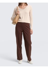 Pull beige à manches longues et col en V, associé à un pantalon large brun et des baskets blanches. Le mannequin tient un sac bandoulière marron clair.