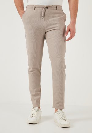 Muž v svetlo béžových slim fit nohaviciach s rukami v vreckách a bielych teniskách, stojaci pred jednofarebným svetlým pozadím.