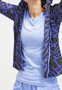 Chaqueta ligera con cremallera de base negra y patrón abstracto morado. Debajo, una camisa de manga corta texturizada de color azul claro.