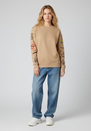 Beige sweatshirt met geribde halslijn, voorzien van kleurrijke graphic en tekst op de mouwen. Gecombineerd met lichtblauwe, loszittende jeans en witte sneakers.