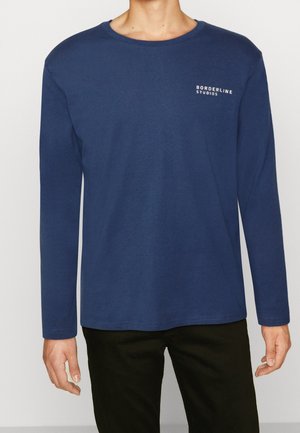 Homme portant une chemise à manches longues bleu marine avec le texte « BORDERLINE STUDIOS » sur la poitrine à gauche, associée à un pantalon noir.