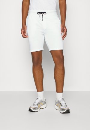 Man draagt witte shorts met trekkoord, witte sokken en grijze sportschoenen, staand tegen een effen lichte achtergrond.