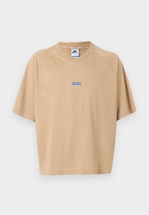 Persoon draagt een beige oversized t-shirt met een blauwe schakellink-stijl nummer "02" op de achterkant, gecombineerd met zwart-witte broek.