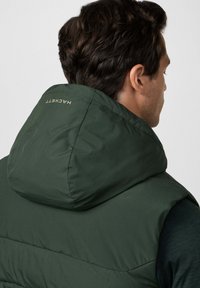 Gilet verde con cappuccio caratterizzato da un design trapuntato, texture liscia e un logo sul retro. Il tessuto appare leggero e resistente all'acqua.