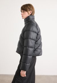 Casaco de puffer em preto com colarinho alto, design matelassé e um corte cropped. Apresenta uma textura suave e detalhe de logotipo nas laterais.