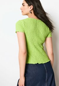 Grünes strukturiertes Oberteil mit kurzen Ärmeln, gewelltem Saum, tailliertem Design, getragen mit einem dunklen Jeansrock, Rückansicht.