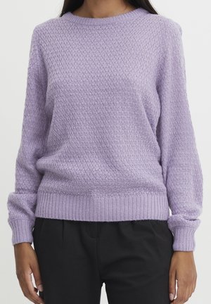 Personne portant un pull en tricot texturé lavande à manches longues et un pantalon noir, debout devant un fond clair uni.