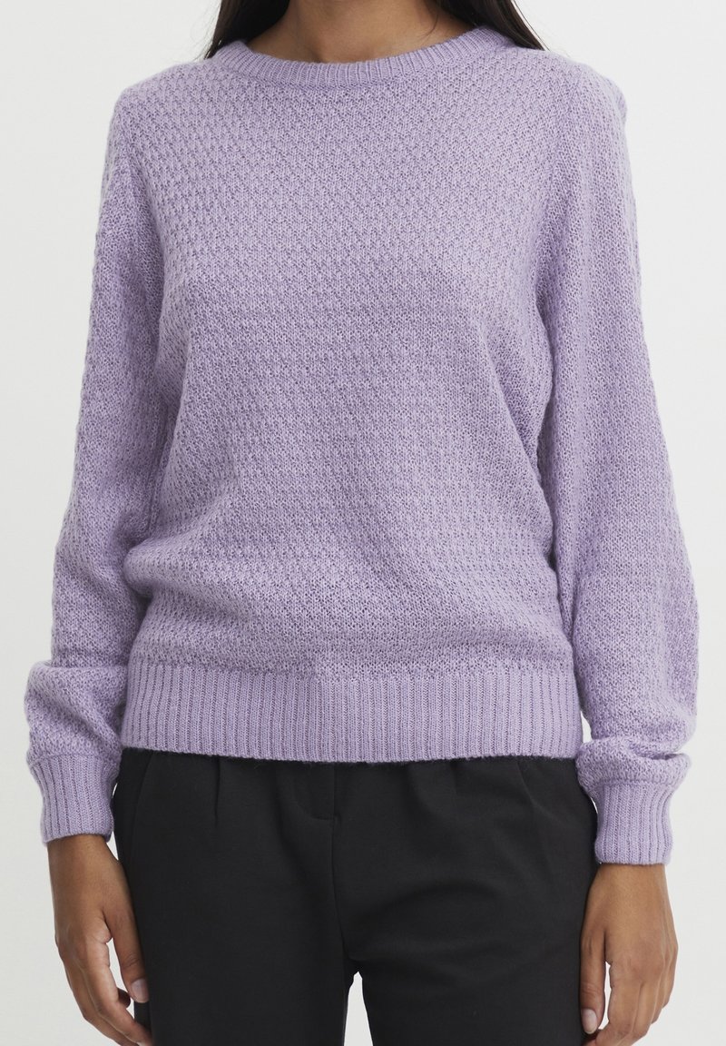 Personne portant un pull en tricot texturé lavande à manches longues et un pantalon noir, debout devant un fond clair uni.
