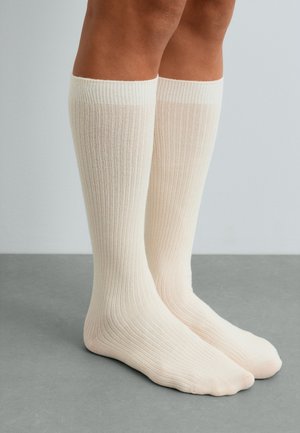 Jambes portant des chaussettes côtelées crème montant jusqu'aux genoux, posées sur une surface lisse grise sur fond uni.