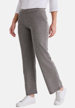 Frau trägt graue, strukturierte Hosen mit geradem Bein, weiße Turnschuhe und ein weißes Langarmshirt, steht vor einem hellen Hintergrund.