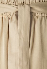 Béžový textilní pásek se zavázanou uzlou. Elastický pas s sklady, vytvářející jemný spad. Hladký, lehký materiál.