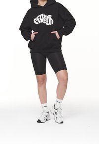 Svart oversized hoodie med vit "Pecador" text, parat med svarta cykelbyxor och vita sneakers med svarta accenter samt loggosockor.