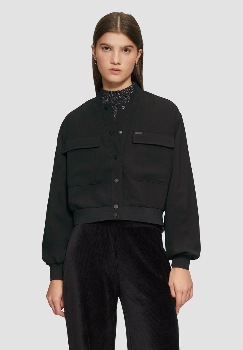 Czarna krótka kurtka z dwoma kieszeniami na piersiach, zapięciem na guziki oraz prążkowanymi mankietami. Wykonana z gładkiego materiału, charakteryzująca się minimalistycznym wzornictwem.