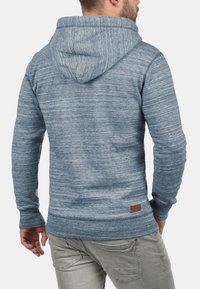 Sudadera azul jaspeada con capucha ajustable, puños acanalados y un acabado texturizado; presenta un pequeño parche de logo de cuero marrón en la parte baja de la espalda.