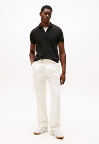 Černé polo tričko, bílé kalhoty a bílé tenisky. Oblečení má klasický střih, hladkou texturu a minimální branding na tričku.