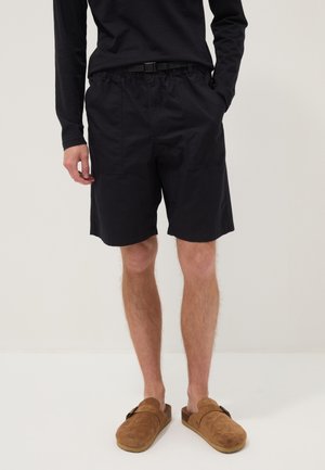 Persona che indossa pantaloncini neri con tasche, una maglia nera a maniche lunghe e scarpe marroni senza lacci, in piedi davanti a uno sfondo semplice.