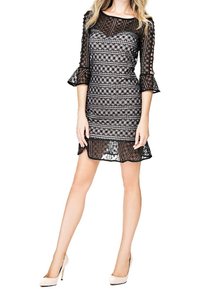 Robe en dentelle noire avec manches longues transparentes, motifs géométriques et poignets évasés. Silhouette ajustée avec détail à volant à l'ourlet, associée à des talons beiges.