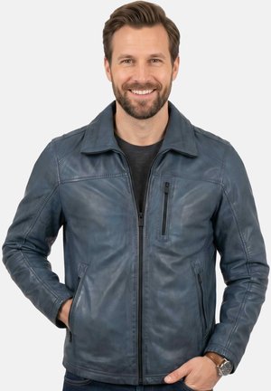 Lächelnder Mann trägt eine blaue Lederjacke mit Reißverschlusstaschen und eine Armbanduhr, steht mit teilweise in den Taschen steckenden Händen.