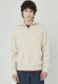 Pull en beige à zip avec une texture côtelée et des manches longues, porté par-dessus une chemise bordeaux, associé à un pantalon à rayures sombres.