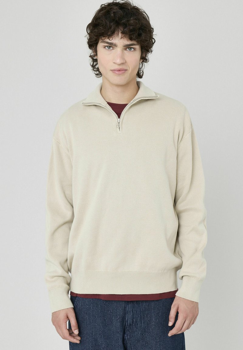 Pull en beige à zip avec une texture côtelée et des manches longues, porté par-dessus une chemise bordeaux, associé à un pantalon à rayures sombres.