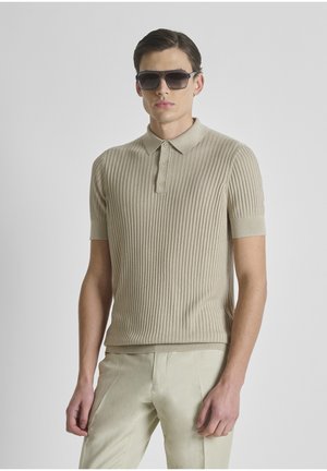 Jonge man met beige geribbelde korte mouwen polo, lichtbeige broek en oversized donkere zonnebril tegen een effen achtergrond.
