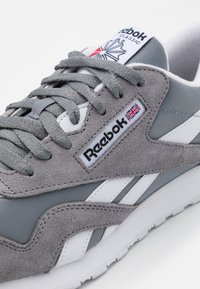 Grå semsket og mesh sneakers med hvite detaljer, som har flate grå snører, et Reebok-logo og en fremtredende polstret tunge.