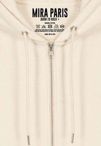 Beige Kapuzenjacke aus Bio-Baumwolle mit Metallreißverschluss, weißen Kordeln und gedrucktem Pflegeetikett innen. Einfaches und funktionales Design.