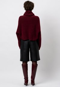 Persoon staat met de rug naar de camera, gekleed in een bordeauxrode coltrui, zwarte leren shorts en bordeauxrode kniehoge laarzen met hakken.