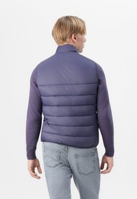 Gilet rembourré violet foncé, avec un col haut et un design matelassé horizontal. Associé à un pull ajusté à manches longues et un jean clair.