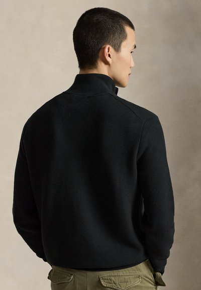 Homme aux cheveux courts noirs, portant un pull noir texturé à col montant et un pantalon vert olive, de dos sur un fond beige.