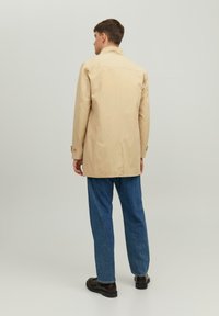 Beige trenchcoat met een rechte snit, rugventilatie en knoopmanchetten, gecombineerd met blauwe denimbroek en zwarte schoenen. Gladde textuur.