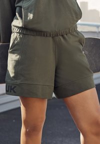 Olive green shorts with an elastic waistband, side pockets, and a black "UK" print on the hem. Made from a soft, textured fabric.
