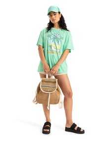 Green oversized t-shirt with palm tree graphic, beige shorts, woven straw backpack with multicolored accents, and black slide sandals.
