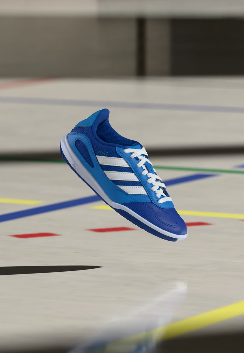 Chaussure de football en salle bleue et blanche avec des lacets, flottant au-dessus d’un sol de gymnase marqué de lignes multicolores.