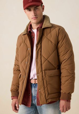 Jonge man die een bruine doorgestikte jas met een corduroy kraag draagt, een rood overhemd, wit T-shirt, blauwe spijkerbroek en een rode pet, staand tegen een beige achtergrond.