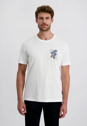 Man in een wit T-shirt met korte mouwen, versierd met abstracte blauwe en bruine vormen en de tekst "pierre cardin" op de borst, staande tegen een witte achtergrond.