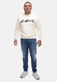 Maglione crema con "St. Moritz" in blu scuro, jeans in denim e scarpe sportive grigie. Il maglione presenta polsini e orlo a coste.