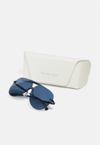 Michael Kors solbriller med mørkeblå linser, en unik aviator-form og en svart stropp, sammen med et hvitt teksturert etui.